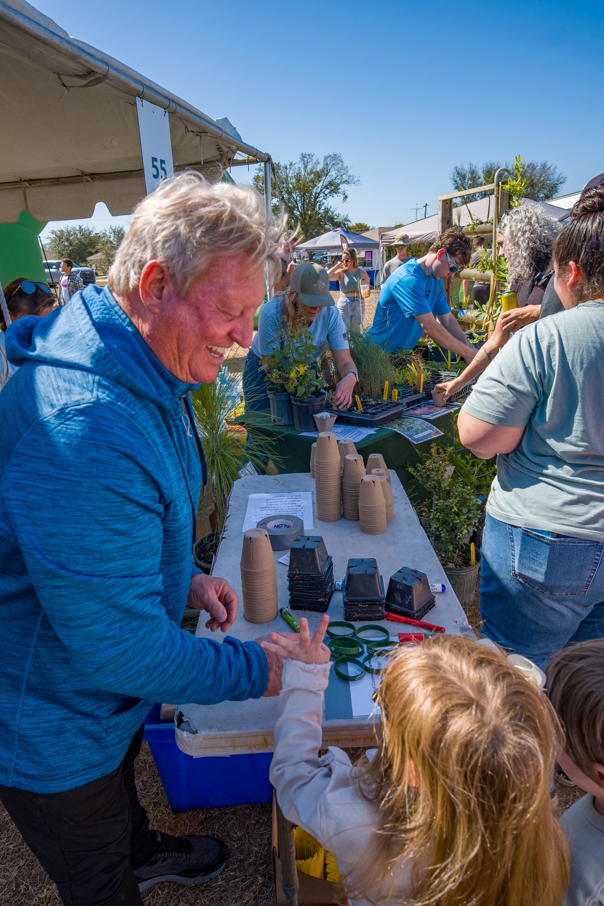 An older man laughs with a young child at a table of small plant pots while volunteers hand out native plants at the Waterways Festival.